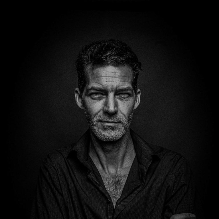 Ein Mann sitzt nachdenklich auf einem Stuhl, gekleidet in dunkler Kleidung und Mütze.